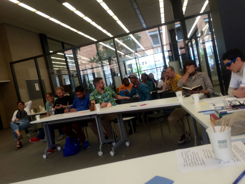 A group of people sit at a semicircle of white tables. They are talking or writing in notebooks.