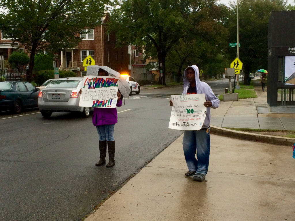 A man in a grey hoodie and a woman in a purple coat with an umbrella stand by an urban street with a grey car passing behind them. The woman holds a sign reading "AFFORDABLE HOUSING NOW!!" The man's sign reads "Only $235 MILLION for housing people can afford, but $700 MILLION to arrest & lock them up? THAT DOESN'T MAKE CENTS! #BalanceTheScales"