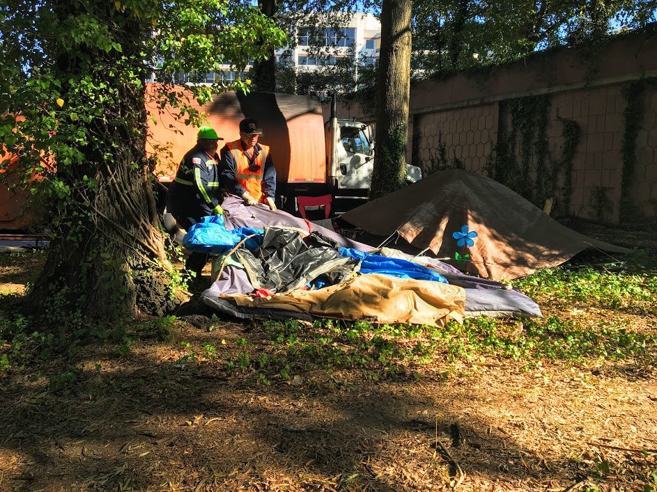 Two city workers drag a tent full of belongings to a garbage can