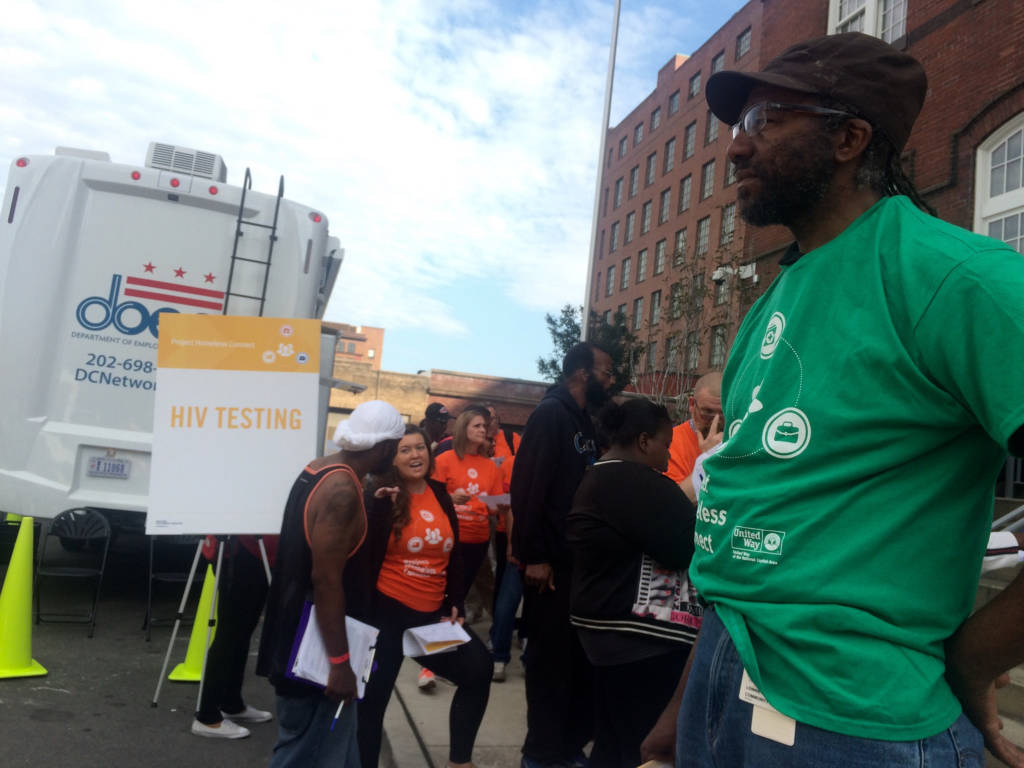 A sidewalk outside the building with volunteers and participants waiting in line. In the left background, there's a white truck and a sign that reads "HIV TESTING". In the right foreground, there's a close up of a volunteer in a green shirt.