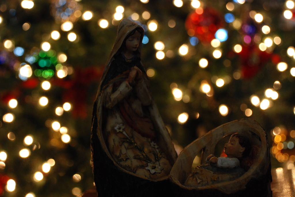 In the foreground, two nativity figures of the Virgin Mary and infant Jesus. In the background is an out-of-focus Christmas tree with string lights.