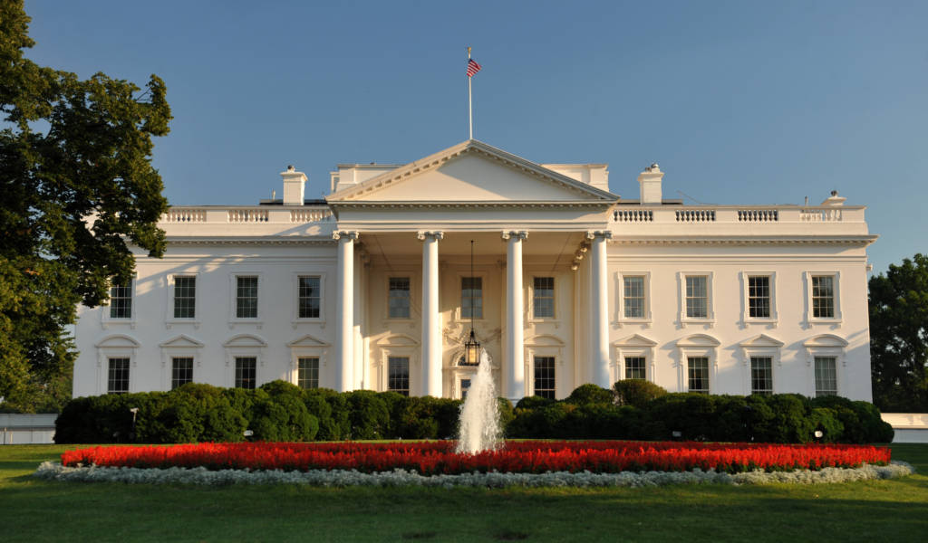 The White House in front of a blue sky with a fountain on the property visible.