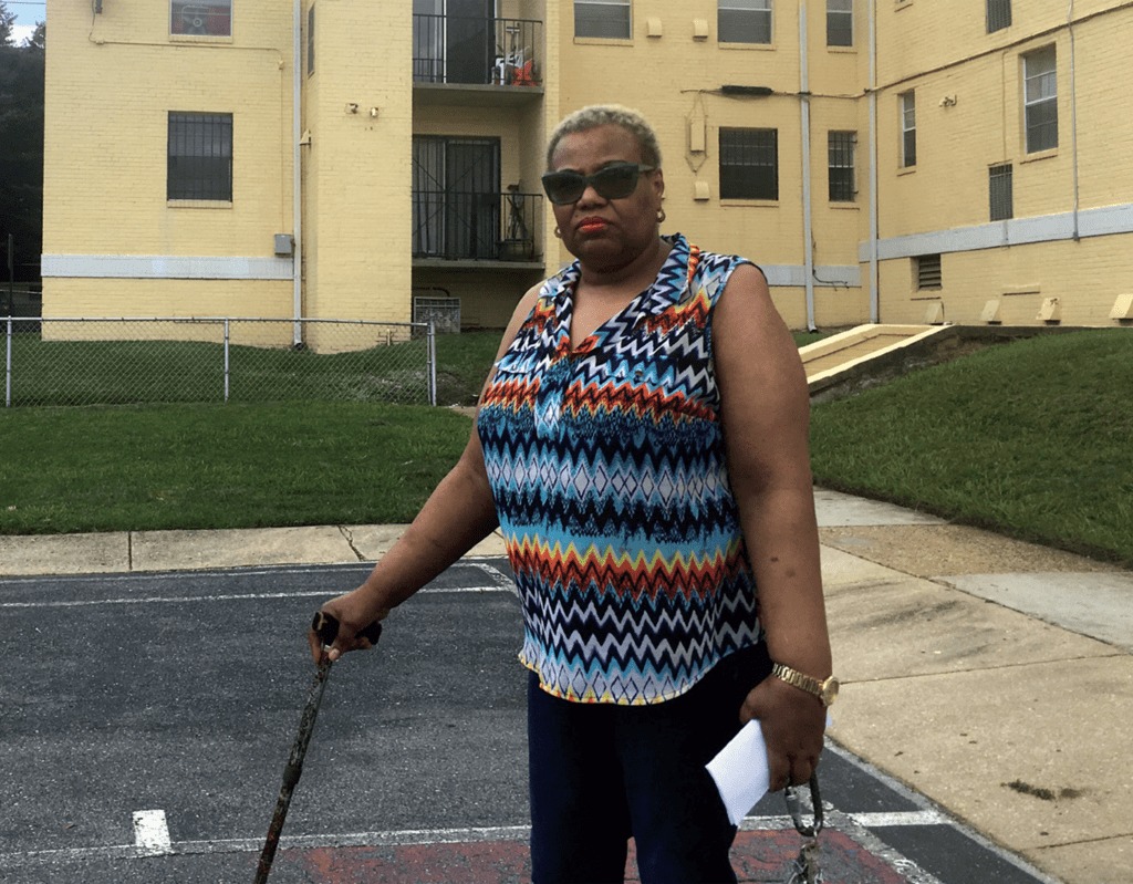 Photo of a woman standing outside of a residential building, using a cane and holding some papers.