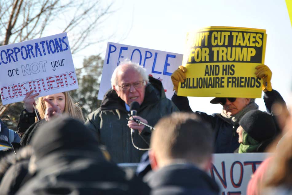 An old man speaks at a podium surrounded by protesters holding signs