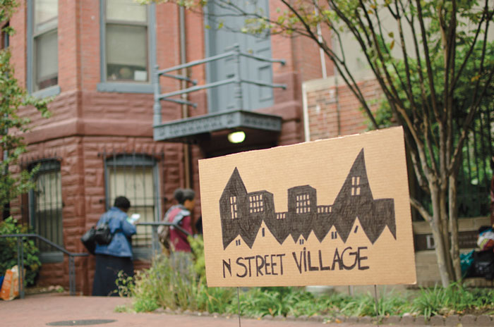 Photo of a sign cardboard sign that reads "N STREET VILLAGE" with a sketch of houses, stuck in the grass outside a home.