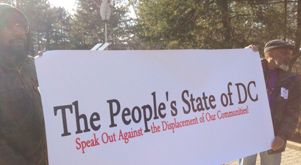 Photo of two people carrying a banner between them that reads: "The People's State of the D.C.: Speak Out Against the Displacement of Our Communities."