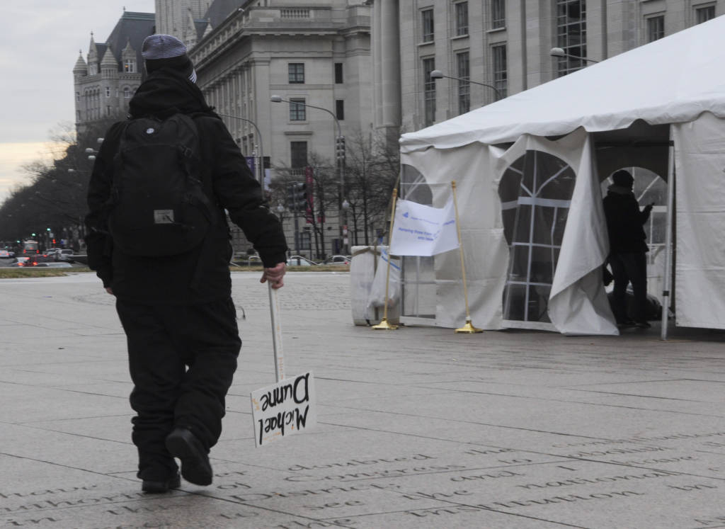 Photo of a white tent on a paved plaza, with a sign in front of it, a man walking towards it and the morning sunrise visible in the sky.