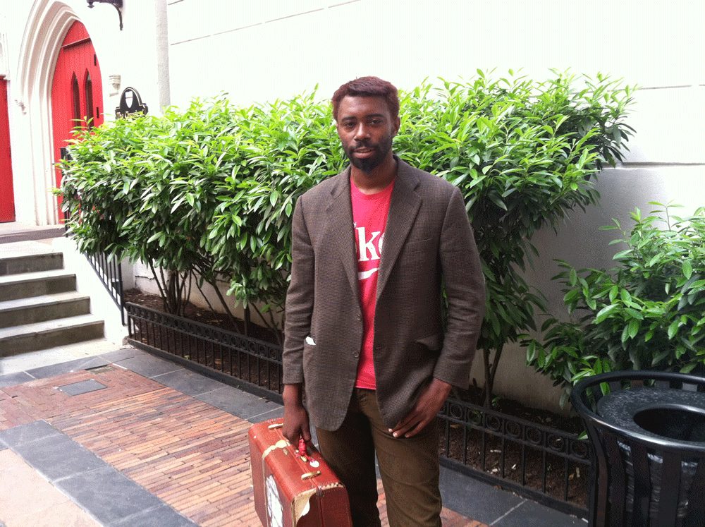 Dele Akerejah stands in a courtyard, wearing a red t-shert with the "Coke" logo under a tweed jacket and carrying a briefcase.