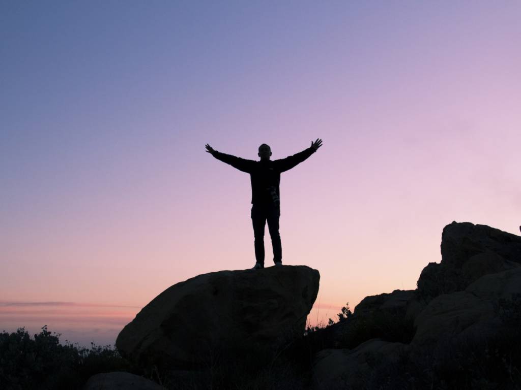 A photograph of a man with arms opened wide, standing on a large rock, looking at the sea.