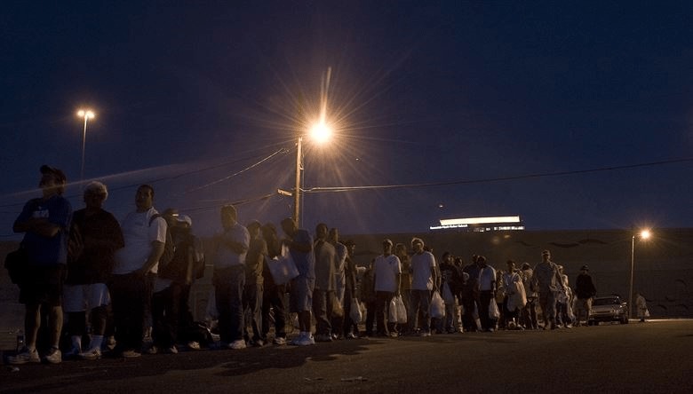 A photograph of a line of people at night, carrying their belongings and waiting to be fed .