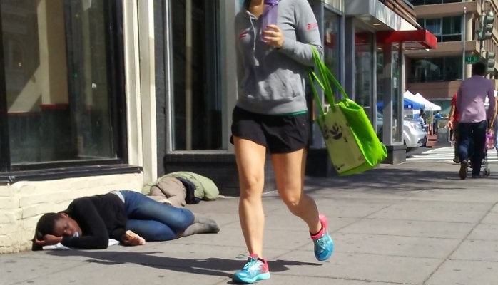 A pedestrian walking past a homeless person laying on the ground.