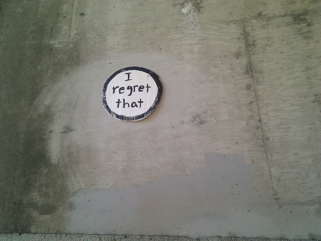 A photograph of a cement wall with a small, circular sign reading "I regret that."