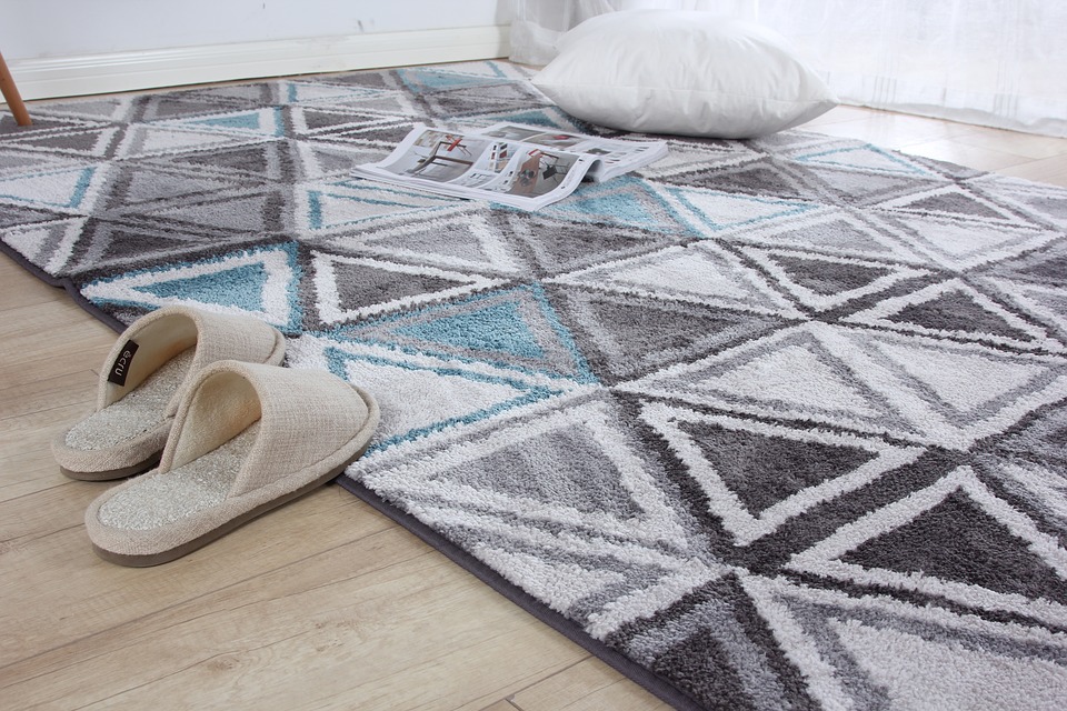 A photograph of a blue, white, and black patterned carpet with a pair of white bedroom slippers sitting nearby.