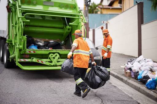 A photograph of two workers in orange jumpsuits loading a green trash truck.
