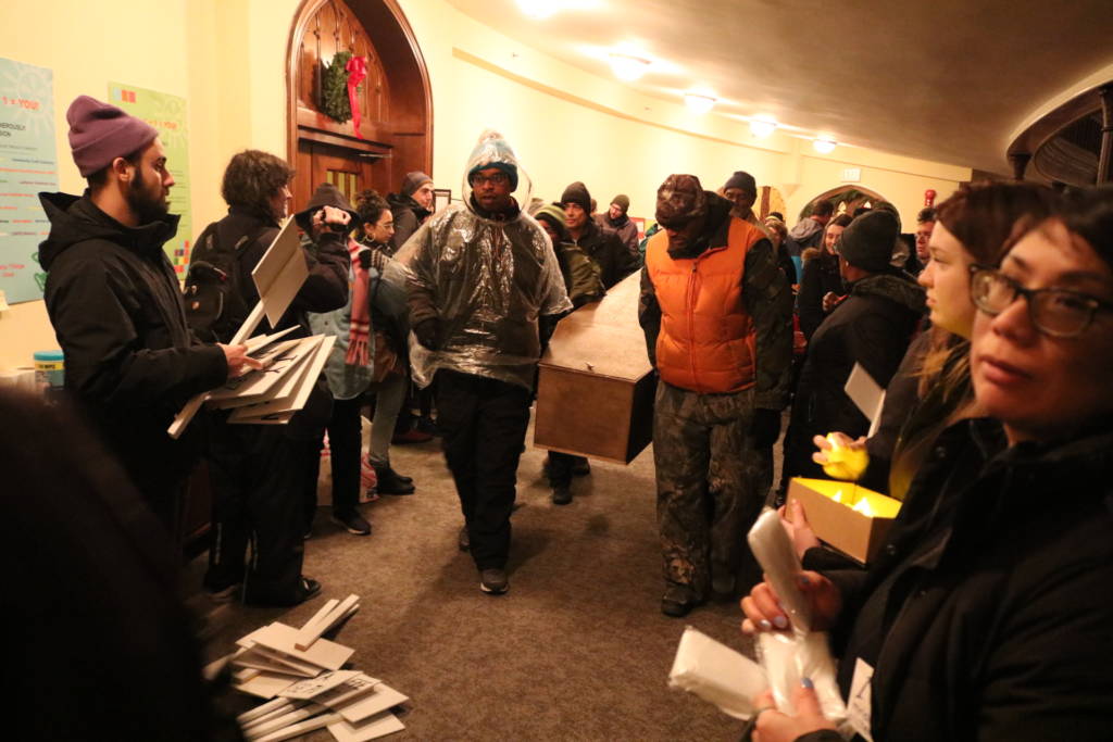 Photo of a coffin being carried through a room People are gathered to each side and signs with names on them are piled on the ground.