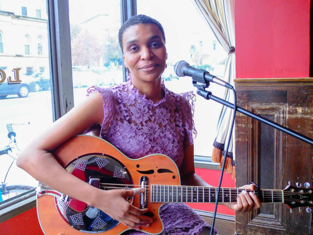 Color photo of a woman in a purple dress sitting in a chair with a guitar in her lap and a microphone stand next to her