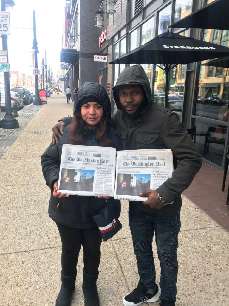 Photo of two people wearing rain jackets and holding newspapers, standing on a sidewalk in front of a Starbucks cafe.