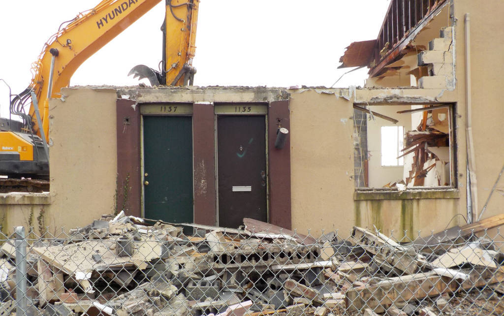 Photo of a residential building facade with two doors. The walls above and to the left of the doors have been ripped away and a yellow construction vehicle is visible behind the wall.