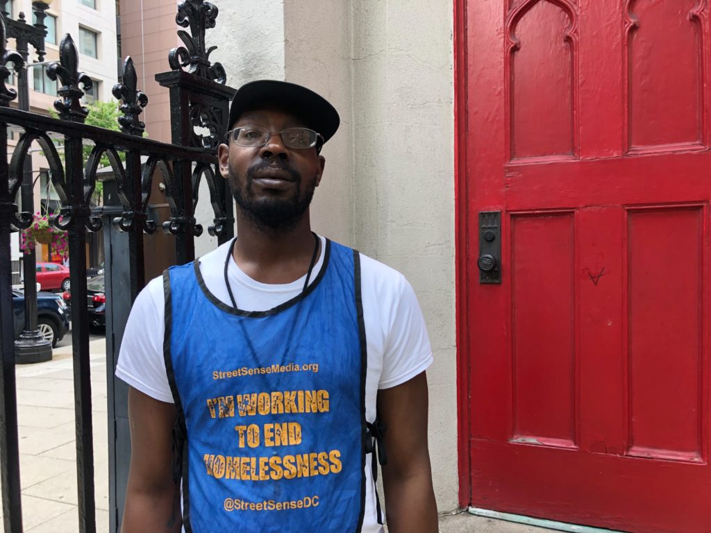 Photo of a man in a blue vest standing by a red door and a black gate.
