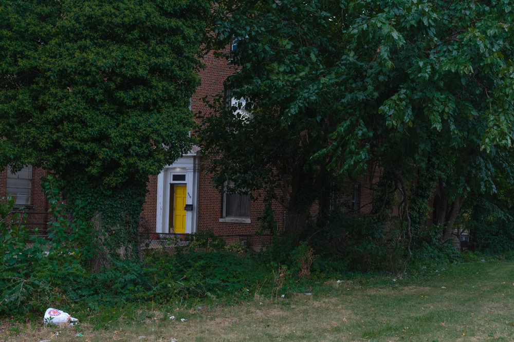 A red brick building with a yellow door and white trim, mostly eclipsed by trees.