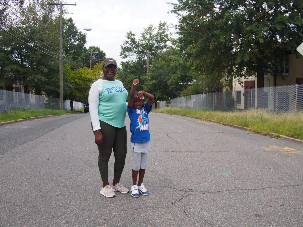 Photo of a woman wearing a teal shirt and a young boy wearing a blue shirt standing in the middle of a paved road with fenced off houses visible behind them.