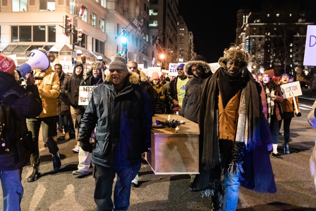 Two African-American male marchers carry a coffin topped with black roses and candles. One marcher holds a megaphone. Other marchers carry signs with the names of those that died.
