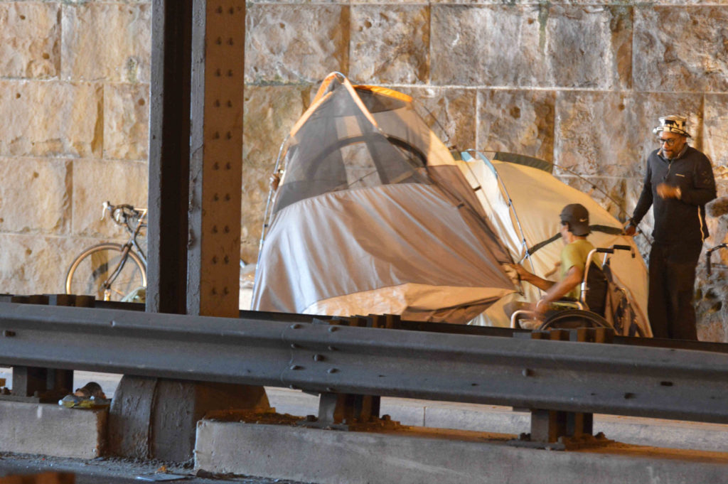 A man in a wheelchair lifts up the edge of a tent in an underpass.