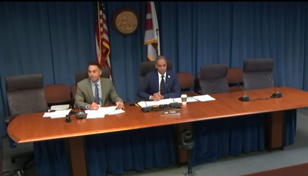 Brandon Todd, dressed in a suit and tie, sits at a long brown desk with papers and a microphone in front of him.