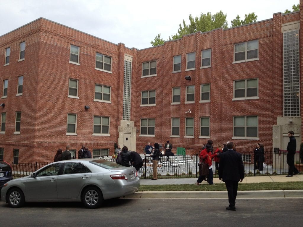 The exterior of a brick apartment building, with some people and cars in front of it.