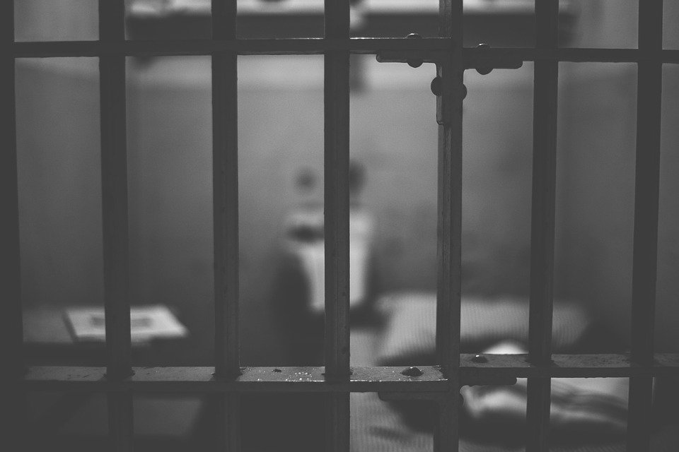 A blurry image of a jail cell. A table, a bed, and a sink are visible.