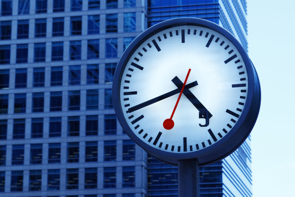 A clock sits in a street in front of an office building.