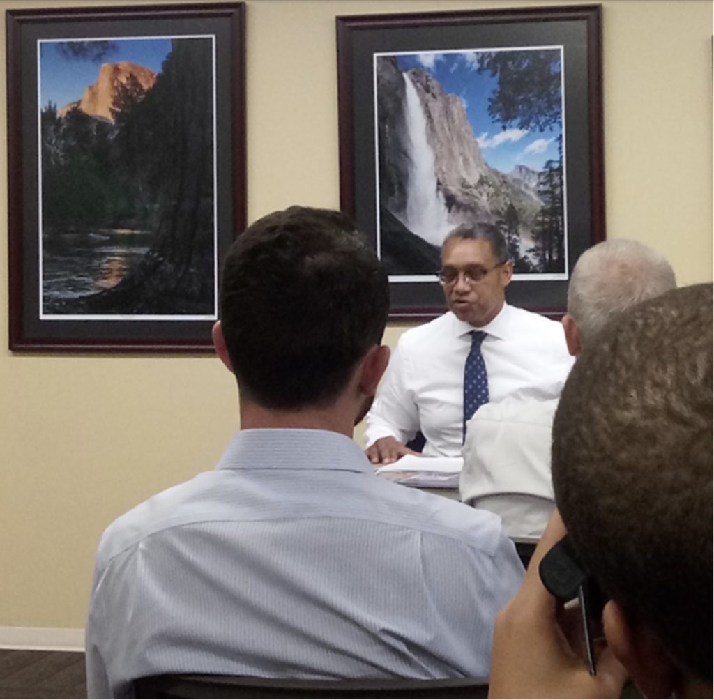 A black man in a white shirt and blue tie sits in front of two posters. One depicts half dome in Yosemite and the other depicts a waterfall.