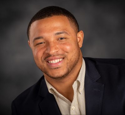 A black man smiling. He wears a white button up shirt without a tie and a black suit jacket