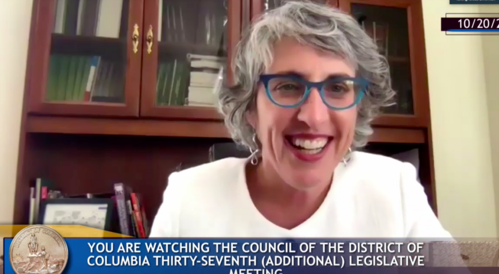 On a video call, a woman with grey hair in a white suit smiles at the camera.