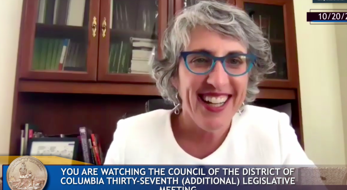 On a video call, a woman with grey hair in a white suit smiles at the camera.