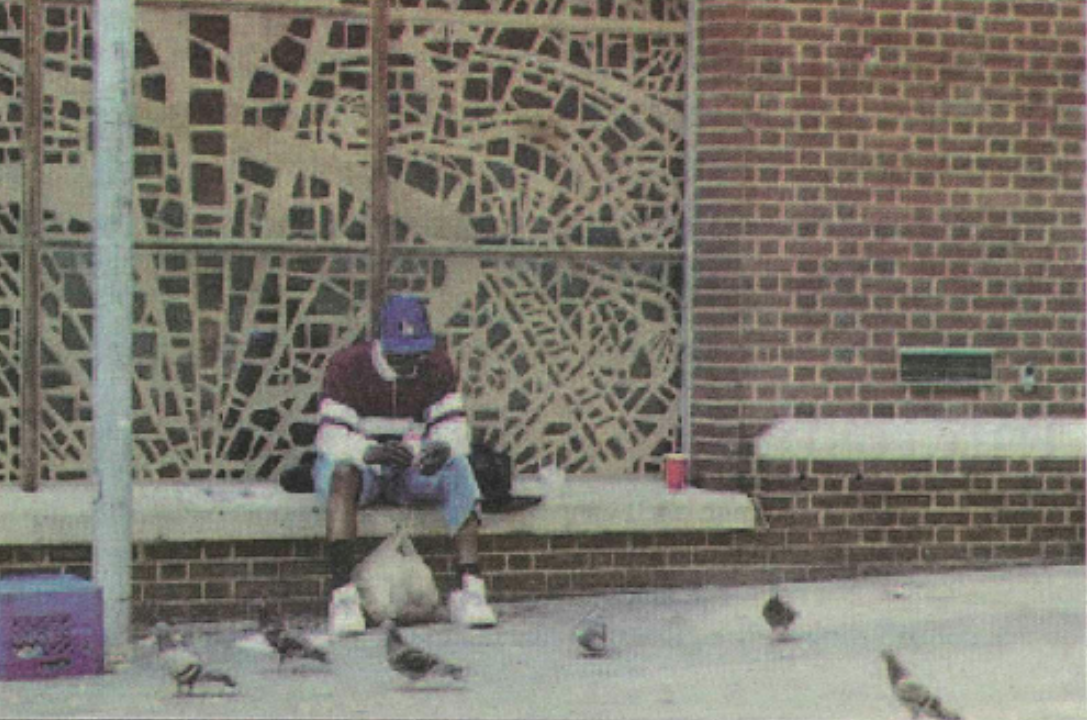 A photo of a man sitting on a bench on the sidewalk.