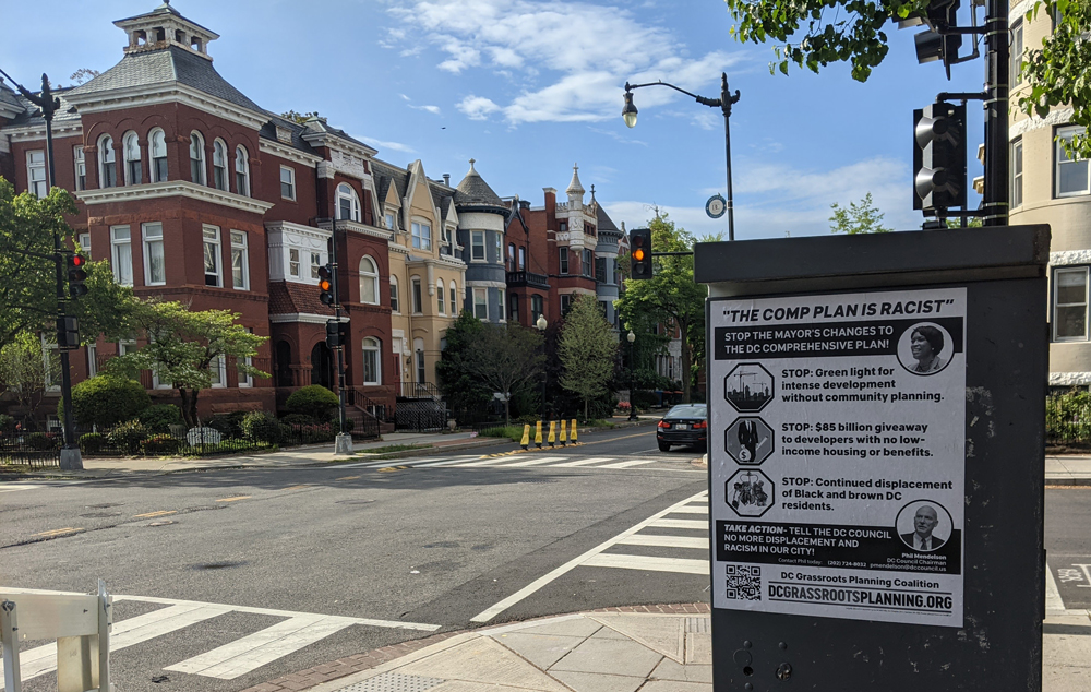 Poster says "The Comp Plan is Racist. Stop the mayor's changes to the DC comprehensive plan! Stop green light for intense development without community planning. Stop $85 billion giveaway to developers with no low-income housing or benefits. Stop continued displacement of Black and brown DC residents. Take Action: Tell the DC Council no more displacement and racism in our city." Row houses are visible behind where it has been tacked up.