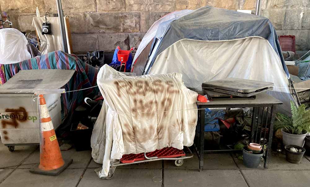 A photo of belongings and encampment of a person experiencing homelessness, which includes a sign asking for help