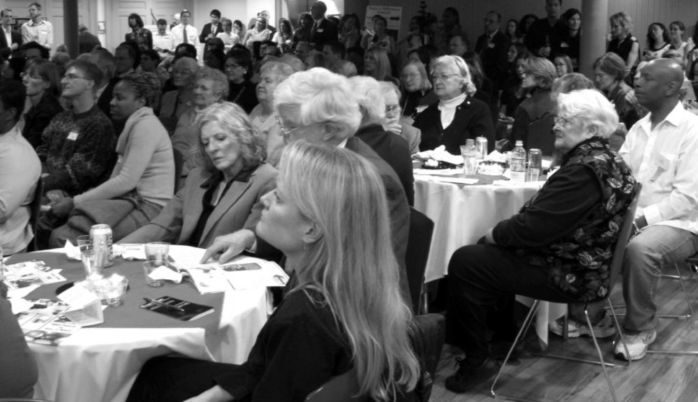 Photo of attendees seated at tables for the event; it is so crowded some attendees must stand in the back