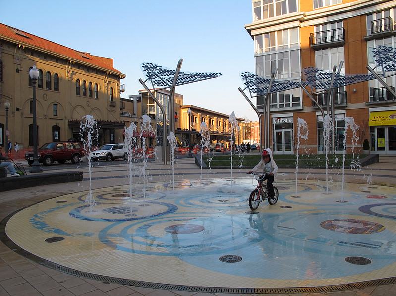 Photo of the public fountain in Columbia Heights, Washington, DC. A young child rides his bike through blasts of water.