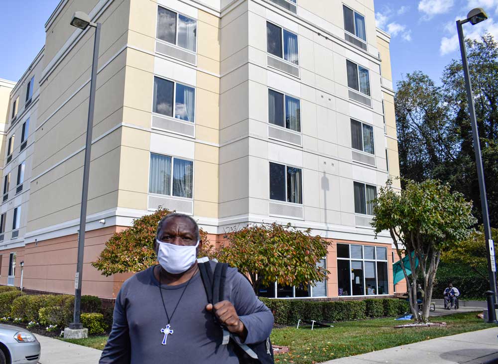 Photo of a man with a backpack, wearing a cross around his neck, standing in front of a hotel.