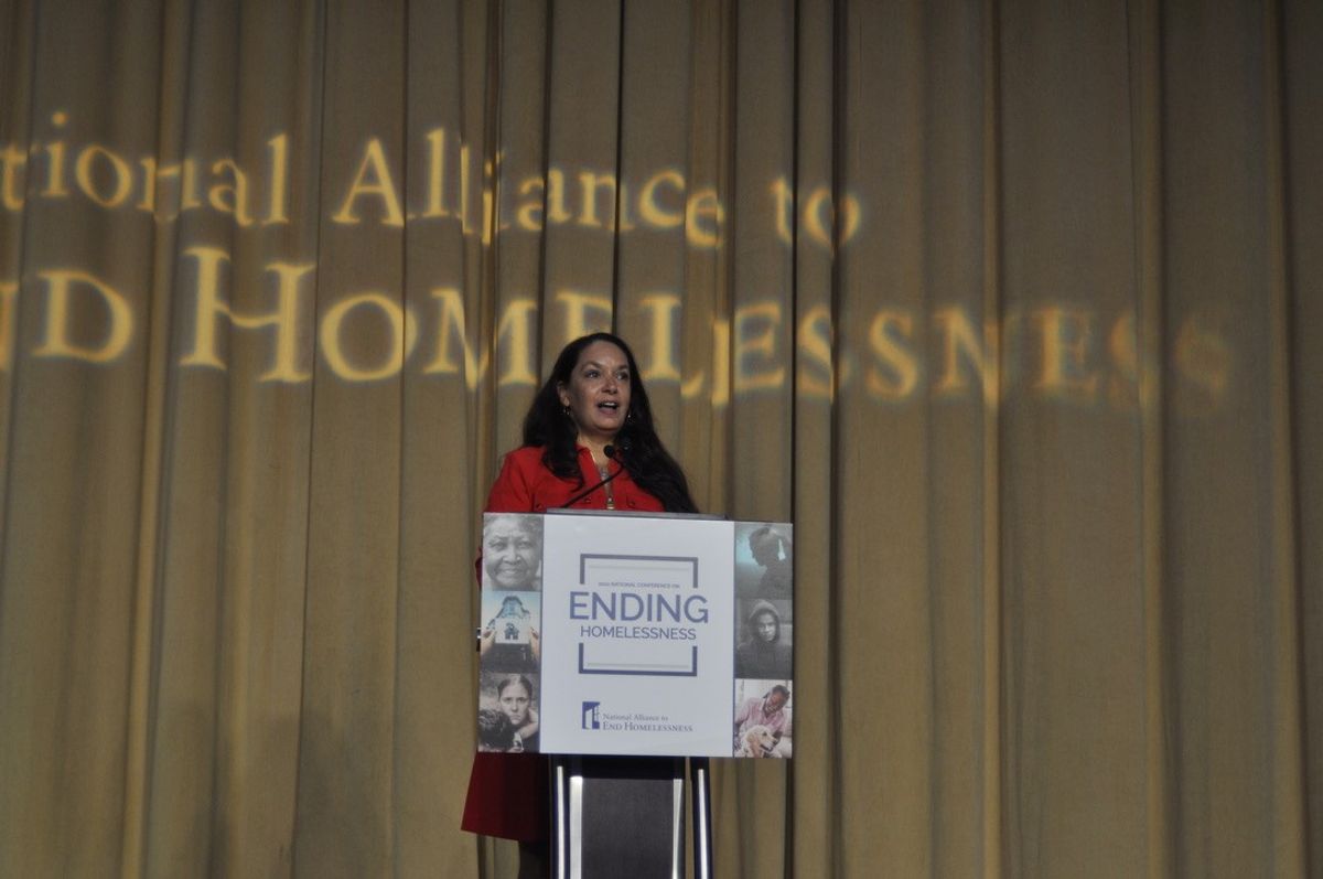 Ann Oliva stands at a podium in front of a gold curtain.