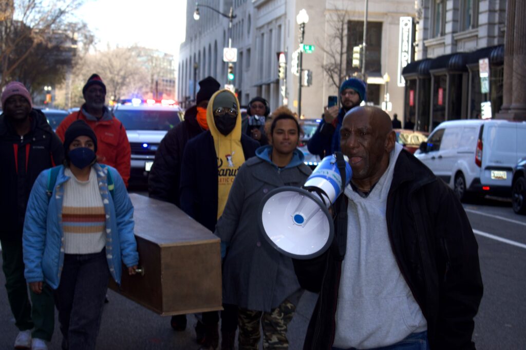 A man yells into a speakerphone as people march on the street behind him.