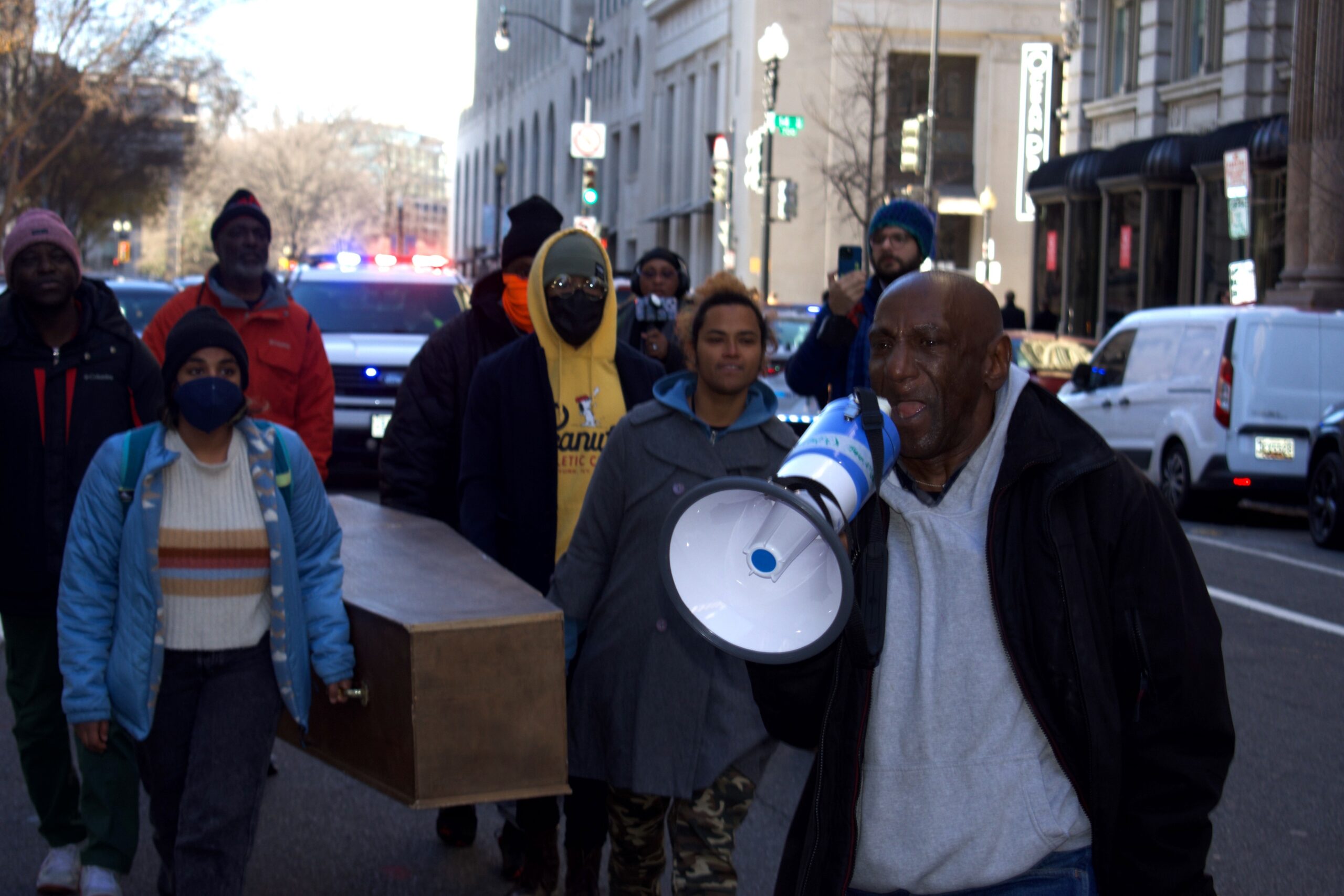 A man yells into a speakerphone as people march on the street behind him.