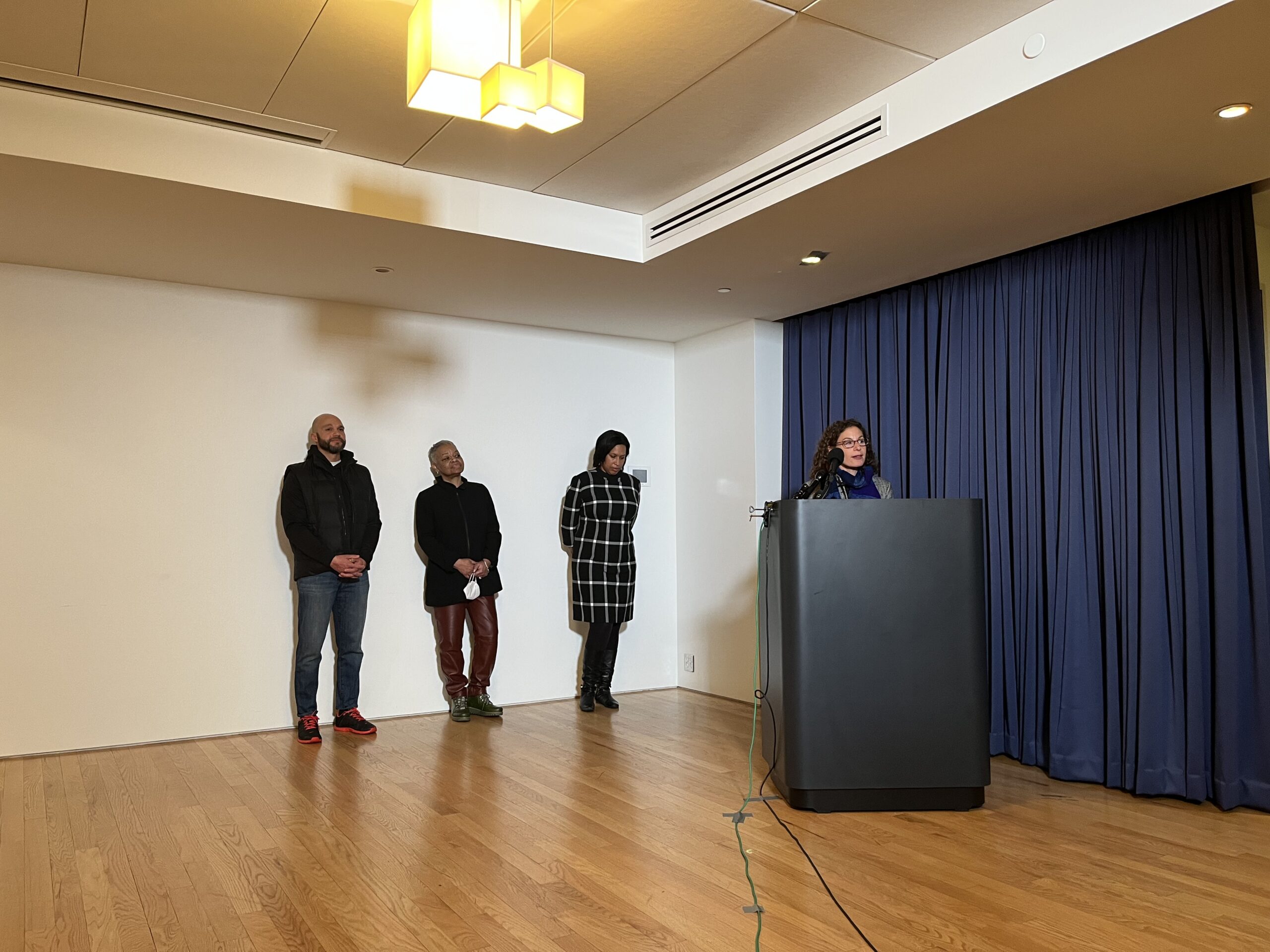 DHS Director Zeilinger speaks from a podium as Councilmember Robert White and Mayor Muriel Bowser look on.