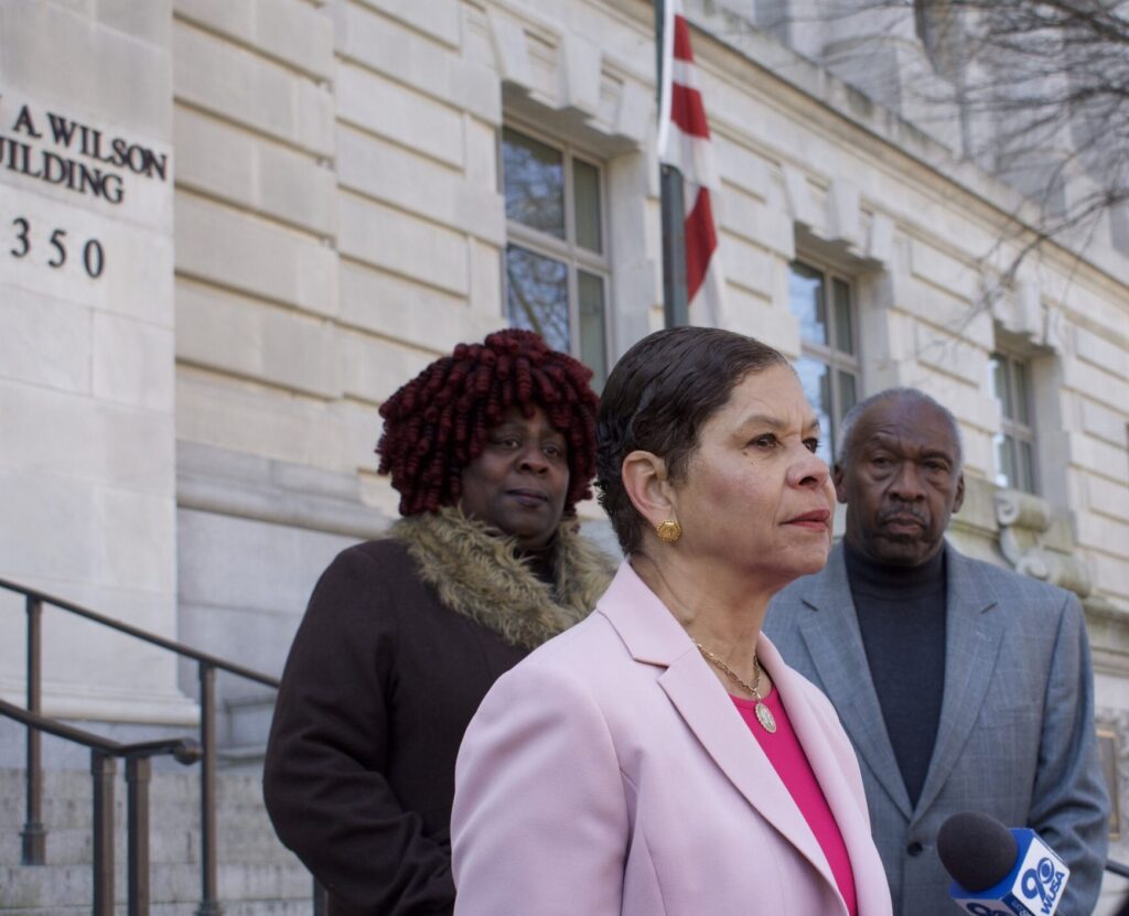 DCHA Director Brenda Donald in a pink suit looks into the distance, with two people behind her.