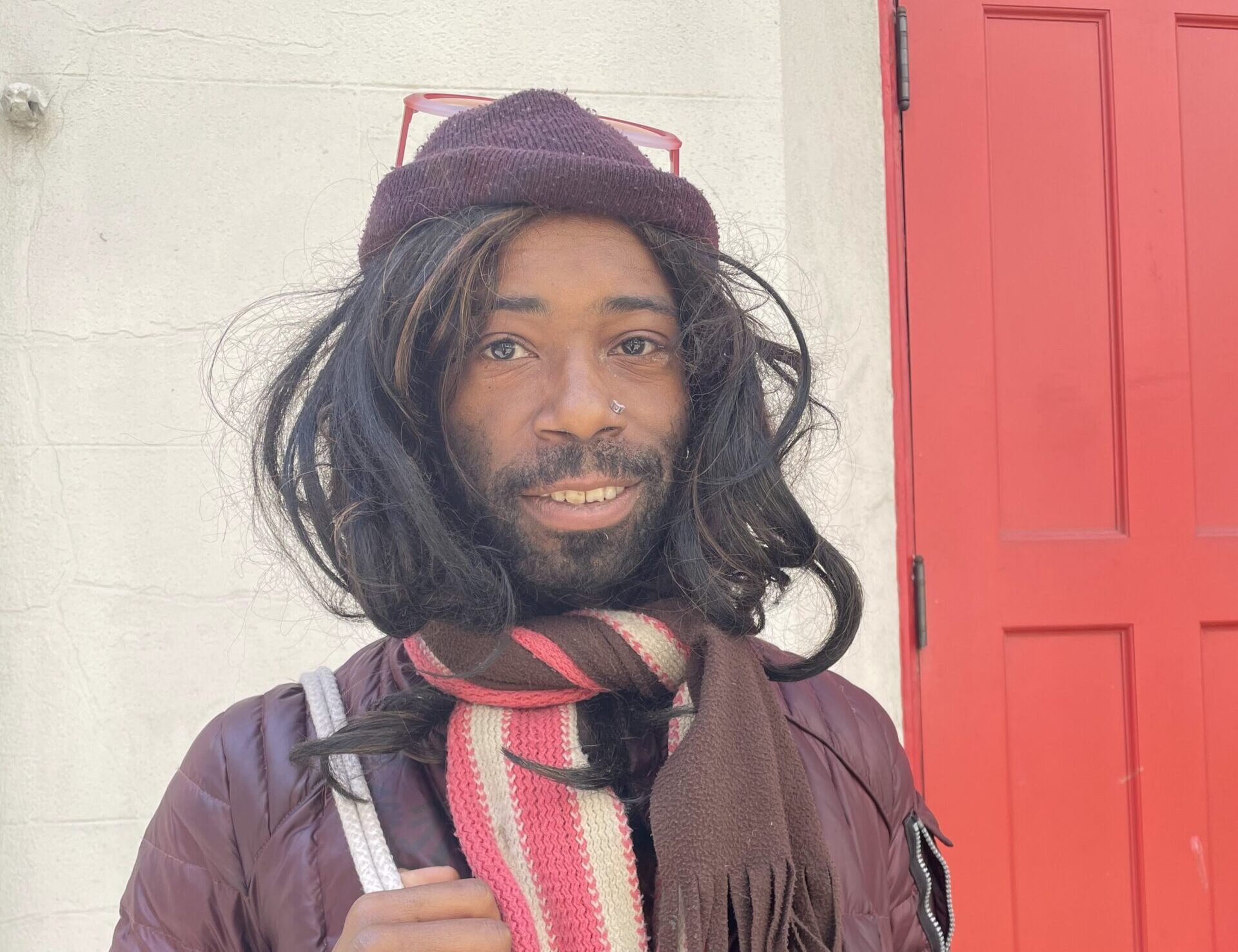 A Black man with long hair, a maroon jacket and hat, and pink scarf and sunglasses smiles at the camera.