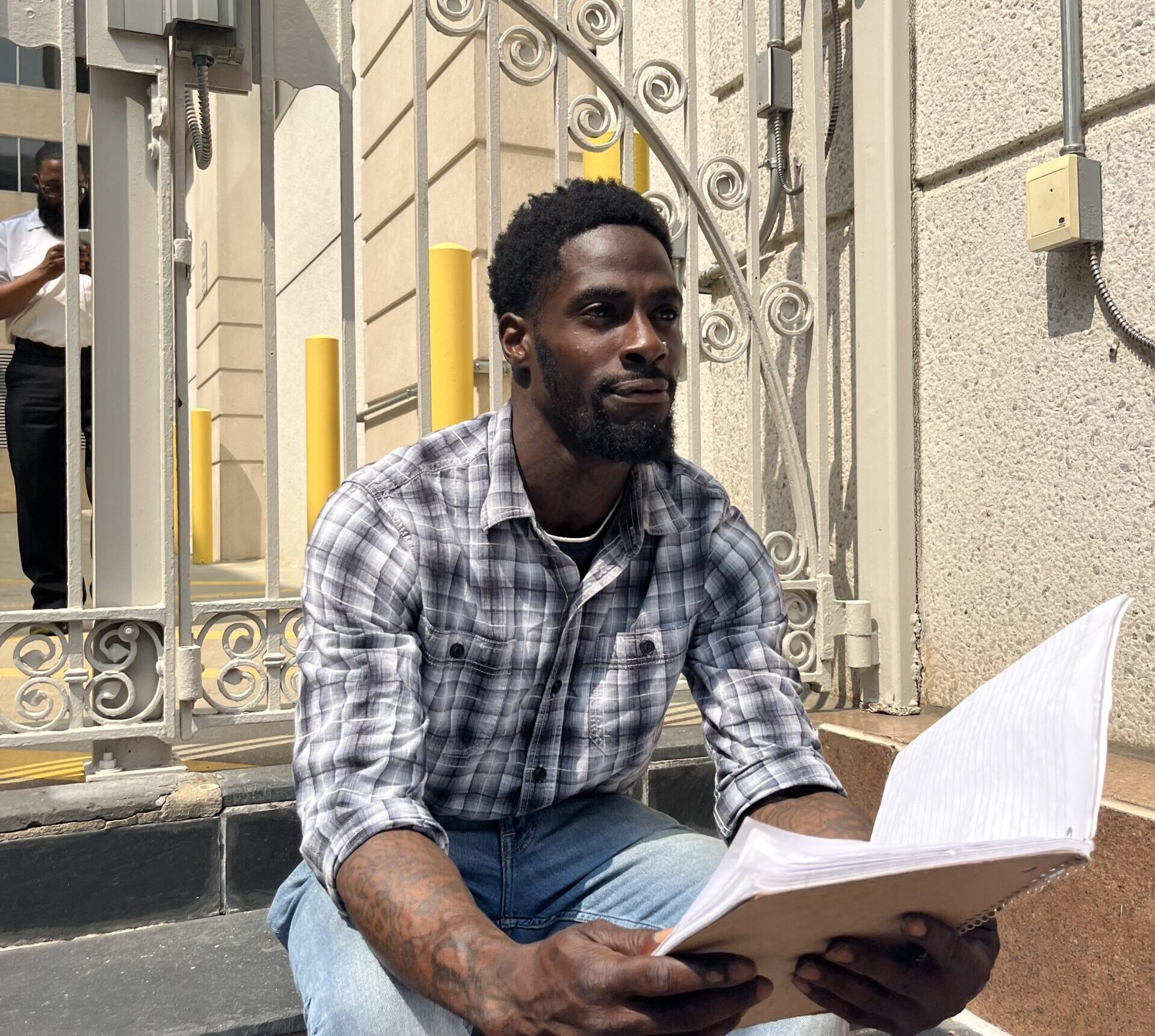A Black man in a plaid shirt and light wash jeans sits on a step and reads from his notebook.