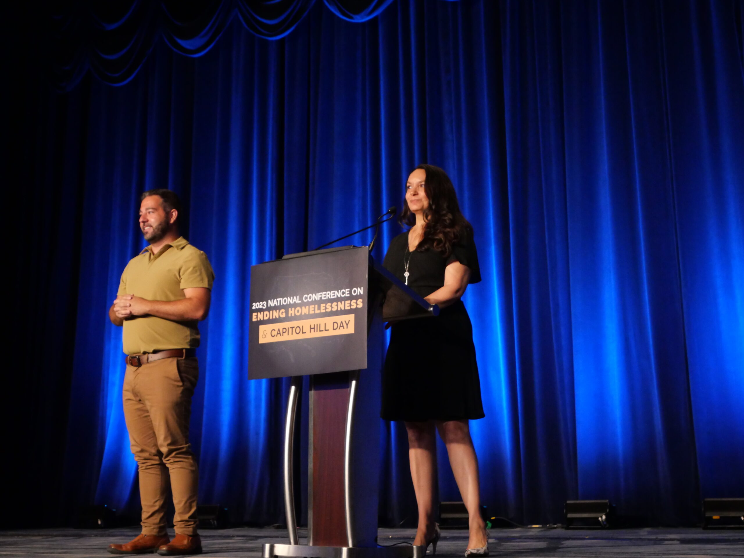A woman in a black dress speaks from a podium, in front of a blue curtain.