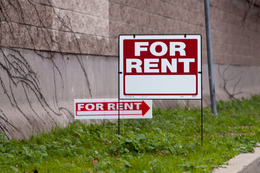 A white and red for rent sign in front of a brick wall. Housing voucher recipients can have trouble renting.